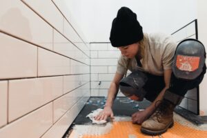 A woman sets tile flooring in a bathroom remodel and wears protective knee pads and work boots
