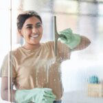 Happy, smile and girl cleaning window with spray bottle and soap or detergent, housekeeper in home